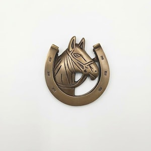 Op de afbeelding: Een bronskleurige metalen riemgesp met een reliëfontwerp van een paardenhoofd in het midden van een hoefijzervorm. Het hoefijzer heeft zichtbare inkepingen voor hoefnagels langs de gebogen rand van het frame.