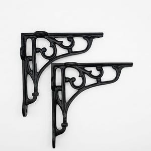 Può includere: Una coppia di reggimensola a forma di L di colore nero con un intricato design a volute. Ogni supporto presenta un bordo superiore piatto per sostenere la mensola e un lato verticale con fori preforati per il fissaggio a parete.