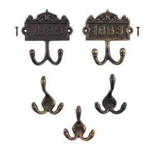 Pode incluir: Um conjunto de cinco ganchos de parede de metal com vários acabamentos em bronze escuro, latão e tons de cobre. Dois ganchos apresentam uma placa retangular gravada com o número 1883 e ornamentos decorativos, acompanhados por dois parafusos.