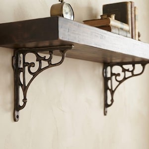 Peut inclure: Une étagère murale en bois brun foncé soutenue par deux supports métalliques ornés de motifs à volutes. L'étagère porte une petite horloge ronde et une pile de trois livres à couverture rigide, présentant une conception robuste.