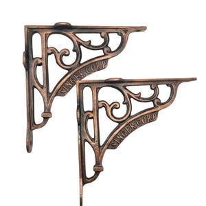 Può includere: Due staffe per mensole a forma di L in metallo color rame scuro con un design ornamentale traforato a volute. Ogni staffa presenta la scritta SINGER CORP in rilievo lungo la barra di supporto diagonale e include fori di montaggio preforati.