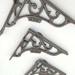 May include: Three dark grey cast iron shelf brackets of varying sizes, each featuring an ornate scrollwork design and the letters GNER embossed on the central horizontal bar. The brackets have pre-drilled holes for mounting to a wall or shelf.