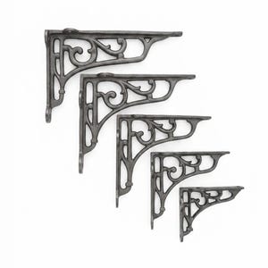 May include: A set of five dark grey metal shelf brackets arranged in a cascading diagonal line. Each bracket features an L-shaped frame with a decorative scrollwork pattern in the corner, including mounting holes at the ends of the vertical and horizontal arms.