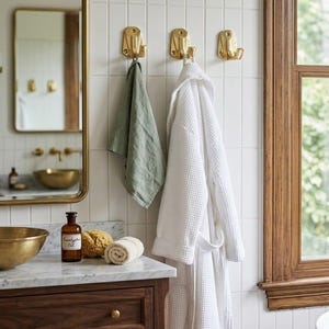 May include: Three polished brass wall hooks mounted on white vertical subway tiles. A sage green linen towel hangs from the left hook, while a white waffle-weave bathrobe hangs from the centre hook. The hooks feature a rectangular base plate.