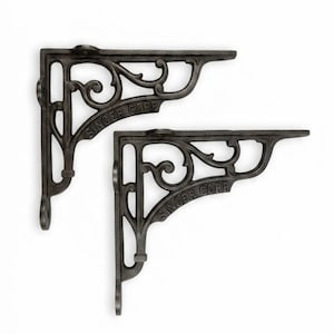 May include: Two dark gray cast iron shelf brackets featuring an ornate scrollwork design and the text SINGER CORP embossed on the diagonal support bar. Each bracket has a vertical and horizontal arm with pre-drilled holes for mounting.