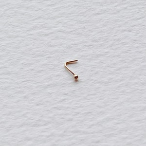 May include: A small, gold-coloured nose ring. It has a curved post and a tiny ball at the end. The jewellery is set against a white background, highlighting its delicate design. A simple, elegant accessory.