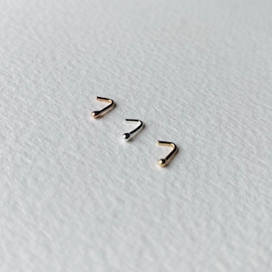 May include: Three nose studs in gold, silver, and rose gold. Each stud has a curved post with a small, round ball at the end. The studs are arranged diagonally against a white, textured background.