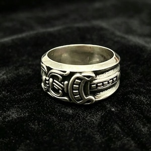 May include: A silver ring with intricate, detailed carvings. The ring features a repeating pattern of curved shapes and rectangular accents around the band. The ring is set against a dark background, highlighting its metallic sheen.
