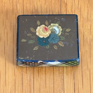 Op de afbeelding: Een kleine, rechthoekige antieke doos met een donkere achtergrond en een bloemenontwerp. De bloemen zijn in tinten rood, geel en blauw. De randen van de doos zijn afgewerkt met een blauw en groen patroon.