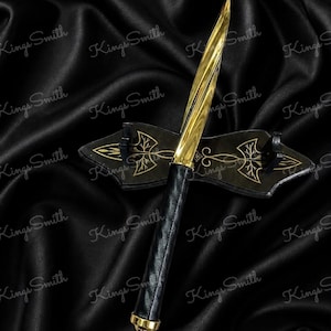 May include: A gold-toned metal dagger with a spiraled blade and a black leather-wrapped handle, featuring a gold pommel at the base. The dagger rests horizontally on a black wooden display stand decorated with gold-colored etched tree motifs.