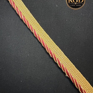 May include: A decorative trim featuring a wide band of metallic gold woven fabric bordered by a thin, twisted cord in a deep magenta red. The trim is positioned diagonally across the frame, accompanied by a circular logo reading RGD LACE SHOP.