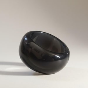 May include: A shiny, black ring with a smooth, curved design. The ring features a thick band and a wide, oval aperture. The surface reflects light, creating highlights and shadows. The ring is a fashion accessory.