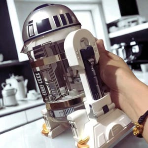 Peut inclure: Une théière en verre argenté et transparent en forme de R2-D2 de Star Wars. La théière a une poignée et une base blanches, avec des accents dorés. Le design comprend des détails noirs et argentés, et est probablement un objet de collection.