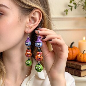 May include: Dangle earrings featuring three stacked charms: a light blue pumpkin wearing a purple witch hat, an orange jack-o'-lantern, and a green jack-o'-lantern. The charms are suspended from silver-toned metal hooks and hardware.