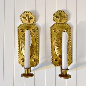 May include: A pair of rectangular brass wall sconces featuring a hammered oval center, a beaded border, and a decorative top finial with a floral motif. Each sconce holds a single white taper candle in a circular brass candle cup at the base.