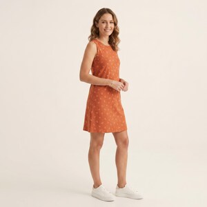 May include: A sleeveless, knee-length dress in a burnt orange hue featuring a subtle, repeating white botanical print. The garment has a round neckline and a straight-cut silhouette, paired with simple white low-top sneakers.