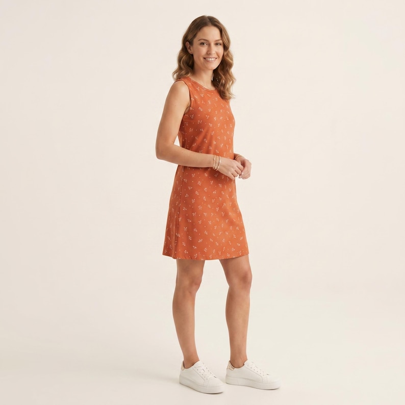 May include: A sleeveless, knee-length dress in a burnt orange hue featuring a subtle, repeating white botanical print. The garment has a round neckline and a straight-cut silhouette, paired with simple white low-top sneakers.