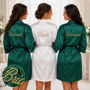Könnte beinhalten: Drei Satin-Morgenmäntel mit langen Ärmeln und Bindegürteln. Zwei Mäntel sind smaragdgrün mit dem Wort Bridesmaid in goldener Schreibschrift auf dem Rücken. Ein Mantel ist weiß mit dem Wort Bride in goldener Schreibschrift auf dem Rücken.