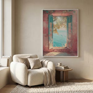 May include: A framed wall art print depicting an open window with teal shutters set into a weathered pink wall, revealing a view of the blue sea and tree branches. The artwork is displayed above a cream-colored armchair and a small wooden side table.