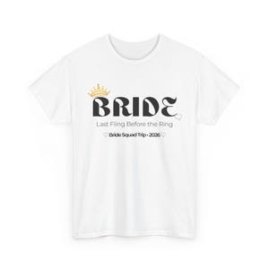 Può includere: T-shirt bianca con la scritta "BRIDE" in un carattere nero stilizzato, sormontata da una corona dorata. Sotto, il testo "Last Fling Before the Ring" e "Bride Squad Trip • 2026" con simboli di cuore.