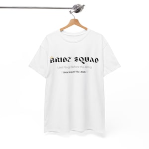 Può includere: T-shirt bianca con la scritta "BRIDE SQUAD" in grassetto, in nero. Sotto, si legge "Last Fling Before the Ring" e "Bride Squad Trip • 2026". La maglietta è appesa a una gruccia di legno.