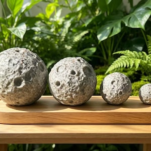 May include: Four spherical objects with a textured, cratered gray surface resembling the moon, arranged in descending order of size on a smooth, light-toned wooden oval tray. The spheres vary in diameter, showcasing a detailed, uneven surface.