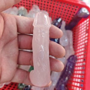 Puede incluir: Una talla con forma de falo hecha de cuarzo rosa pálido y translúcido. El objeto presenta una superficie lisa y pulida con vetas internas naturales y una cabeza redondeada, mostrando la textura cristalina del material de piedra.