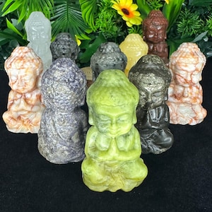 May include: A collection of small Buddha figurines carved from various stones, including lime green, dark grey, purple, white, and reddish-brown minerals. Each figure is depicted in a seated meditation pose with hands pressed together in prayer.