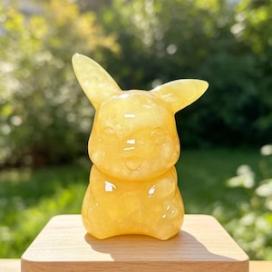 May include: A small, translucent yellow gemstone carving shaped like a stylised creature with two pointed ears, a rounded head, and a smooth, polished surface. The figure is positioned on a light-coloured, rectangular wooden base with a flat top.