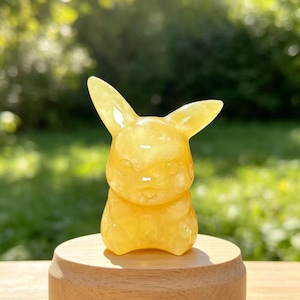May include: A yellow translucent carved stone figurine shaped like a creature with long pointed ears and a small smiling face. The figurine is positioned on a smooth, round, light-coloured wooden base that has a flat top and curved sides.