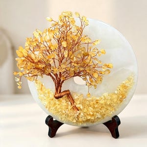 May include: A decorative sculpture featuring a wire tree with yellow crystal leaves mounted on a circular white stone disc. The base of the tree is surrounded by loose yellow crystal chips, and the entire piece rests on a dark wooden stand.