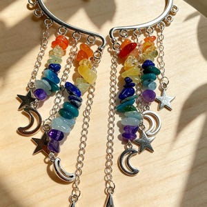 Puede incluir: Un par de pendientes tipo ear cuff plateados con cadenas colgantes adornadas con esquirlas de piedras preciosas multicolores en degradado arcoíris. Cada pendiente incluye pequeños dijes de metal con forma de estrellas y lunas crecientes.