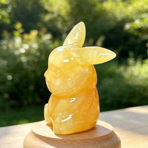 May include: A translucent yellow, polished stone carving in the shape of a seated rabbit with long ears, positioned on a smooth, round, light-coloured wooden base. The figurine features a smooth, glossy surface that reflects light.