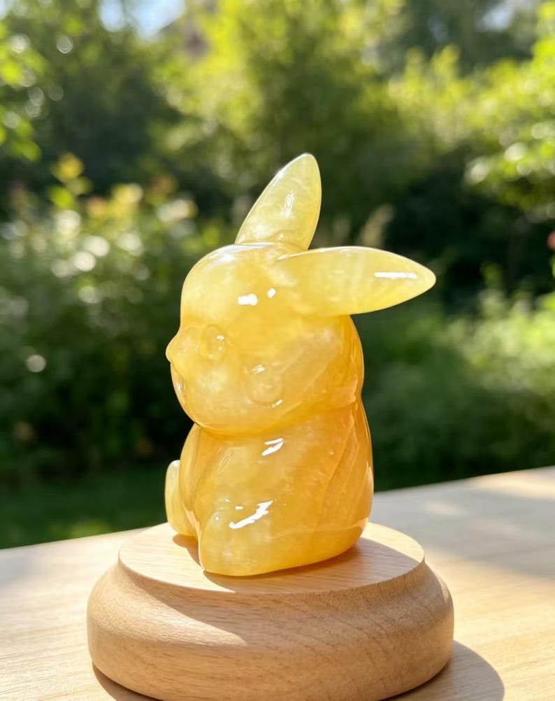 May include: A translucent yellow, polished stone carving in the shape of a seated rabbit with long ears, positioned on a smooth, round, light-coloured wooden base. The figurine features a smooth, glossy surface that reflects light.