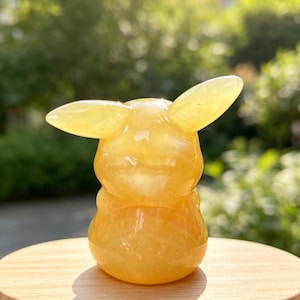 May include: A small, smooth, yellow gemstone carving shaped like a stylised creature with two prominent, pointed ears extending from the sides of its head. The translucent, polished mineral has a rounded body and a glossy, reflective surface.
