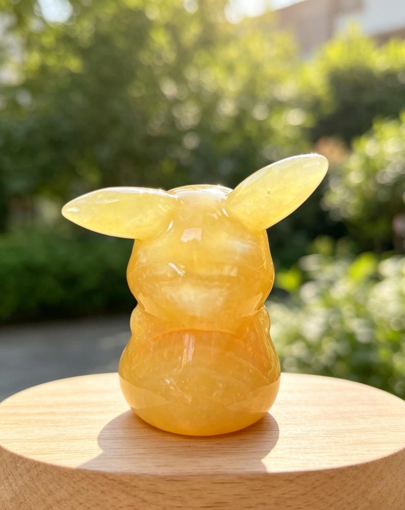 May include: A small, smooth, yellow gemstone carving shaped like a stylised creature with two prominent, pointed ears extending from the sides of its head. The translucent, polished mineral has a rounded body and a glossy, reflective surface.