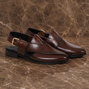 Puede incluir: Un par de sandalias de cuero marrón con correa trasera y hebilla dorada. Las sandalias tienen un tacón bajo y una parte superior de cuero texturizado. Los zapatos se exhiben sobre una superficie de mármol marrón.