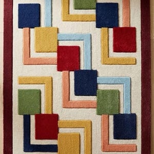May include: A rectangular area rug featuring a cream-coloured background with a deep burgundy border. The surface displays a repeating geometric pattern of raised, tufted squares and L-shaped lines in shades of navy blue, red, yellow, green, and peach.