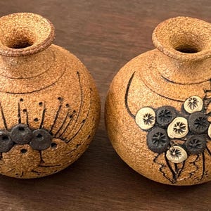 May include: Two small, round, tan-colored ceramic vases with a textured, speckled surface. Each vase features a narrow neck and a flared rim. One vase displays a dark, incised floral pattern, while the other features a cluster of dark berries.