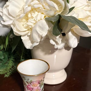 May include: A small, decorative porcelain vase with a floral design featuring pink roses and gold trim. The vase is set on a dark wooden surface, with a white vase of large white flowers in the background.