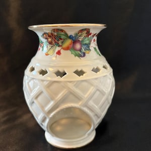 May include: A white ceramic oil warmer featuring a lattice-textured body with a circular cutout at the base. The upper rim is decorated with a colorful botanical pattern of holly leaves, berries, and fruit, finished with a thin gold-tone trim.