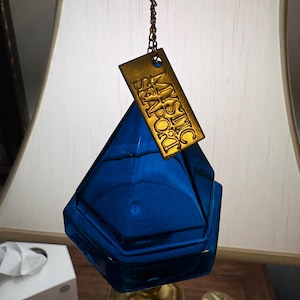 May include: A faceted blue glass ornament with a geometric diamond shape, suspended by a gold-toned metal chain. Attached to the chain is a rectangular gold-colored metal tag featuring the embossed text MYSTIC SEAPORT in capital letters.