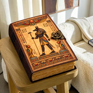 Puede incluir: Un diario rectangular con cubierta color canela que presenta una ilustración de la deidad egipcia Anubis sosteniendo un bastón, flanqueado por dos figuras de aves. La cubierta incluye bordes decorativos, jeroglíficos y un cierre de metal bronce.