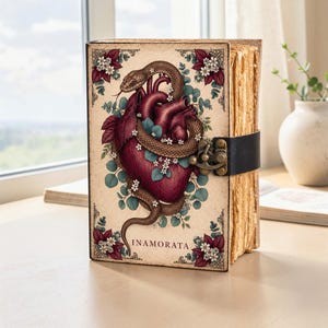 Può includere: Un libro con copertina rigida color crema che presenta un'illustrazione centrale di un cuore anatomico rosso scuro avvolto da un serpente marrone, circondato da piccoli fiori bianchi e foglie verde acqua. La parola INAMORATA è stampata in basso.