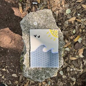 Op de afbeelding: Een rechthoekige stoffen patch met een geborduurd ontwerp van een gele zon, twee zwarte vogels en een blauw golfpatroon met witte geschulpte details op een lichtbeige achtergrond, afgewerkt met een witte stikrand.