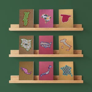 Op de afbeelding: Negen rechthoekige notitieboekjes gerangschikt op drie houten planken. Elk notitieboekje heeft een uitgesneden kaart van een land of continent op de kaft, met diverse patronen zoals bloemen, geometrische vormen en landschappen in kleuren.