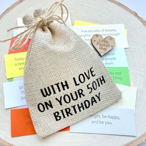 May include: A beige burlap gift bag with the text "WITH LOVE ON YOUR 50TH BIRTHDAY" sits on a wooden slice. A small wooden heart with "Little Pocket Hug" is next to the bag. Colorful notes are scattered around.