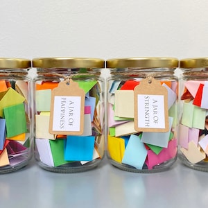 Op de afbeelding: Vier glazen potten met gouden deksels gevuld met kleurrijke papieren vierkanten. Elke pot heeft een bruin papieren etiket met de tekst "A Jar of" en een ander woord: "Friendship", "Happiness", "Strength" en "Positivity".
