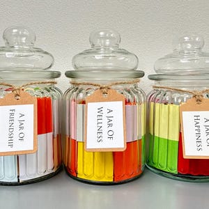 May include: Three glass jars with lids, each containing colorful rolled-up papers. The jars are labeled "A Jar Of Friendship", "A Jar Of Wellness", and "A Jar Of Happiness". Each jar is tied with twine and has a small tag.