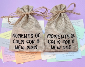 May include: Two burlap bags with the text "Moments of Calm for a New Mum" and "Moments of Calm for a New Dad" printed on them. The bags are filled with small colorful cards.